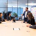 Meeting with six business people sitting around a board table, with whiteboard. A man standing in grey suit with arms open and talking
