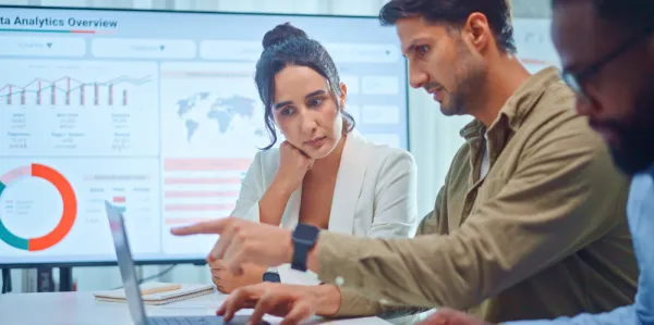 Woman and man sitting at desk with computer and large screen in the background.