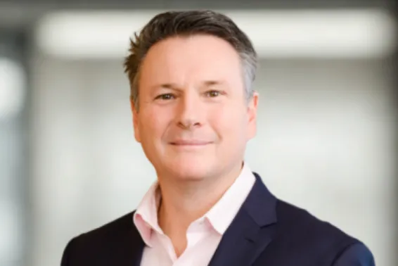 Man with brown hair looking at camera, wearing a pink shirt and black business jacket. Photo from shoulders up.