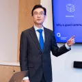 Business man in grey suit, white shirt and blue tile standing in front of a presentation and pointing to content on the wall.