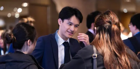 Man in suit with dark hair leaning forward with a hand raised smiling and talking to two females