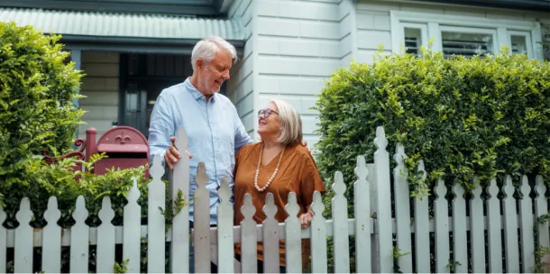 Senior couple embracing while standing near fence in their front yard.