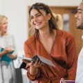 Women with mustard coloured silky shirt and laptop standing near a man and learning forward smiling at someone else