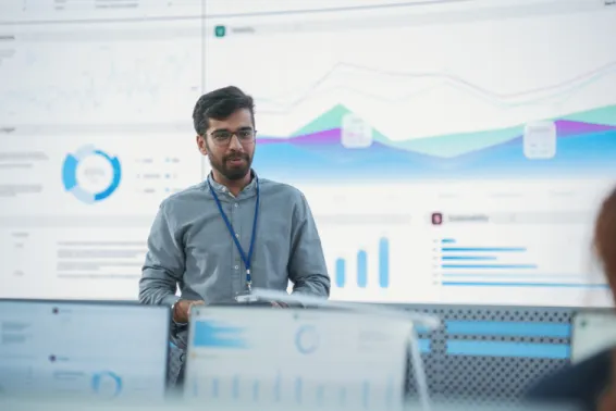 A man talking in front of a large screen with data analysis to co-workers, on their laptops with their back turned to the camera. 