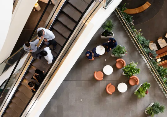 Overshot of a business foyer that captures people on an escalator and others having coffee. 