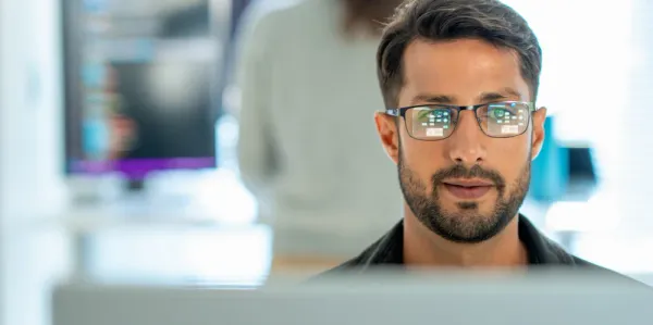 Man in glasses typing at a computer. A lady is typing at another computer in the background