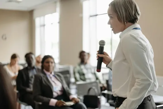 Woman with short grey hair holding microphone speaking to an audience at an event