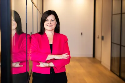 Woman smiling, standing in office hallway, arms folded and bright pink jacket.