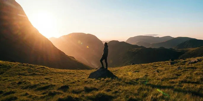 Person standing on a hill beneath mountains at sunrise