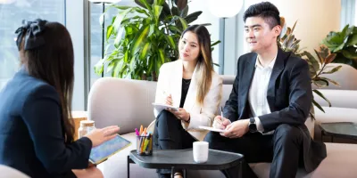 A man and a woman sitting on a business lounge, with pen and paper in hand taking notes, talking to another woman dressed in a dark blue suit.