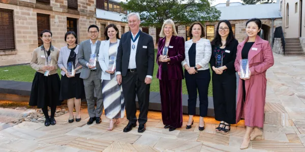 Group of nine people, men and women in business dress, holding awards and standing in the courtyard surrounded by sandstone buildings