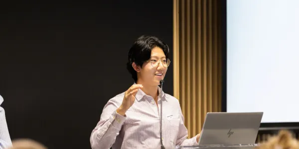 Man with dark hair and glasses in business shirt standing at a lectern smiling with arm raised