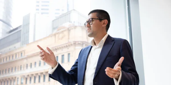 Man standing by large office window with sandstone building in background. Man in blue business suit with white shirt open neck, arms outstretched, smiling and talking