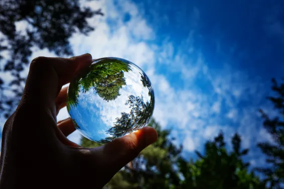 Close up of hand holding glass ball that is reflecting the tress and blue sky background. All Actuaries Summit 2026.