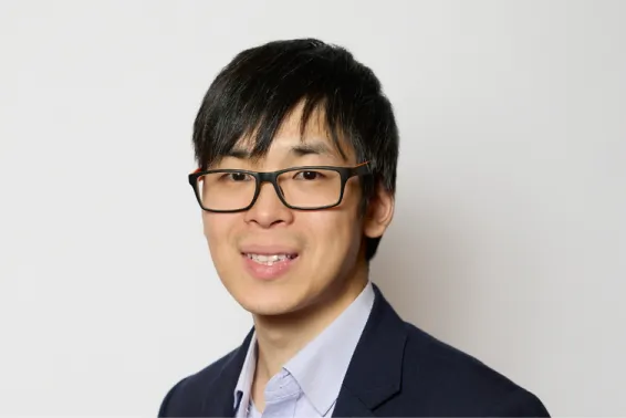 Business man smiling at camera with black hair, black rimmed glasses, and open shirt and black suit