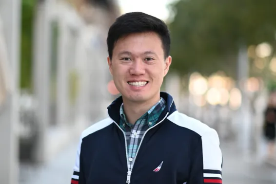 Male looking at camera smiling, with short black hair, and green checked shirt and sports jacket standing in street.