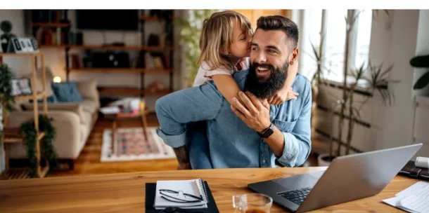 Father working on laptop, with daughter giving him a hug