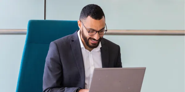 Man sitting on bright blue lounge chair, leaning forward over a laptop and smiling
