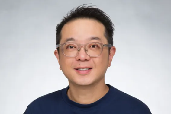 Male with see opaque glasses and dark hair looking at camera. With navy top.