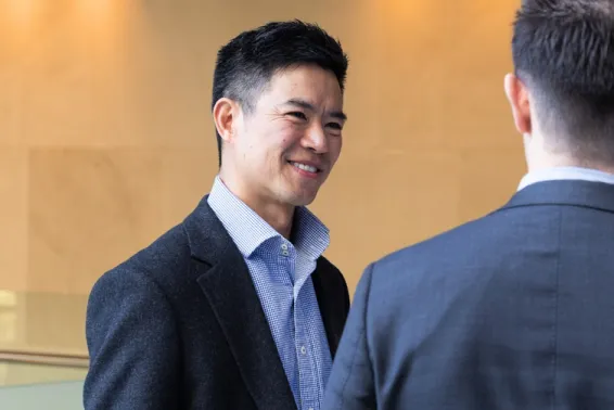 Male dark hair in dark blue suit and open blue shirt smiling and at another male with back to camera