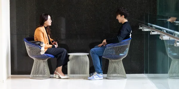 TWO people facing eachother, sitting on chair in a foyer and chatting, leaning forward