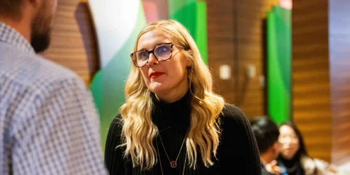 Woman in black polo and long blonde hair, glasses in a work meeting area looking to another person