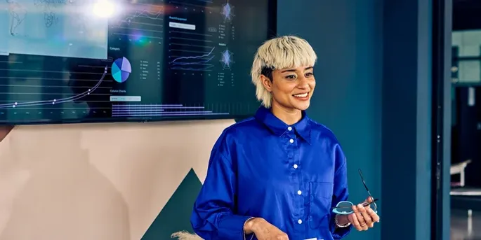 Lady with blonde hair holding glasses giving a presentation in front of a screen with lots of data.