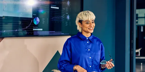 Lady with blonde hair holding glasses giving a presentation in front of a screen with lots of data.