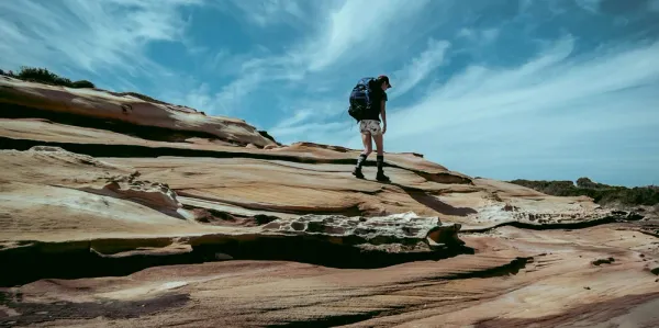 Solo hiker wearing backpack walking up a big rock with blue sky in background.