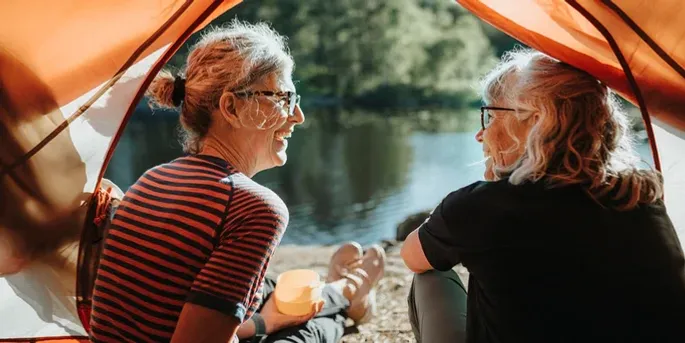 Two older women with grey hair sitting by a riverbank relaxing and having lunch.