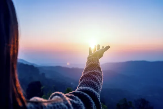 Inspiring image of woman with outstretched, upturned hand to the horizon with sun rising, and mauve skies.