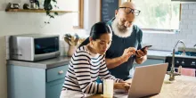 A mature couple using a laptop and phone in the kitchen at home.