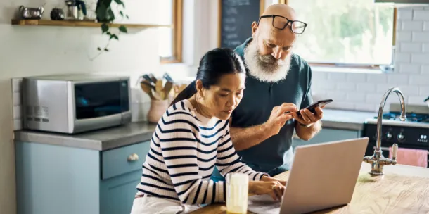 A mature couple using a laptop and phone in the kitchen at home.