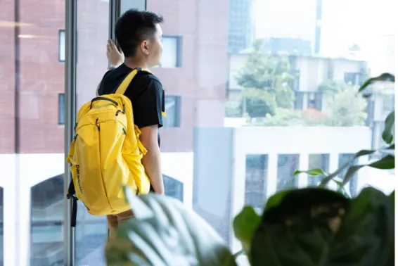 Young man in black tshirt looking out an office window with a yellow backpack over shoulder