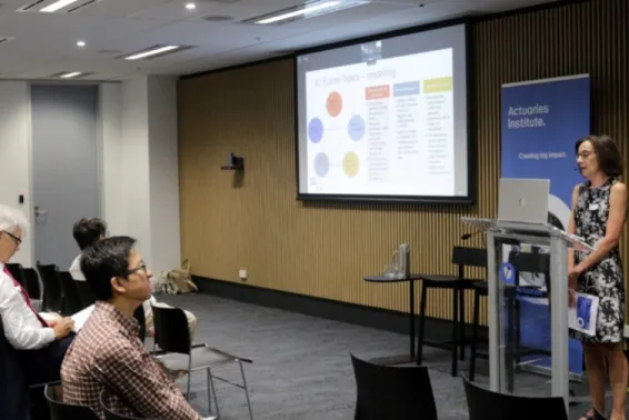 woman stands at lectern in front of group of people sitting.