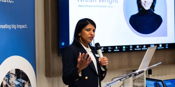 Female with black hair and holding microphone standing in from of screen and banner speaking