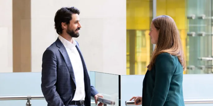 Man in dark business suit leaning in bannister talking to women in a green jacket