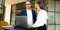 A professional man and woman stand looking at laptop.