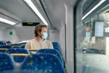 A male passenger sits on an empty train. He is looking out of the window. The Caucasian male is wearing a face mask due to the covid-19 pandemic. The man is commuting to the office as he is an essential worker.