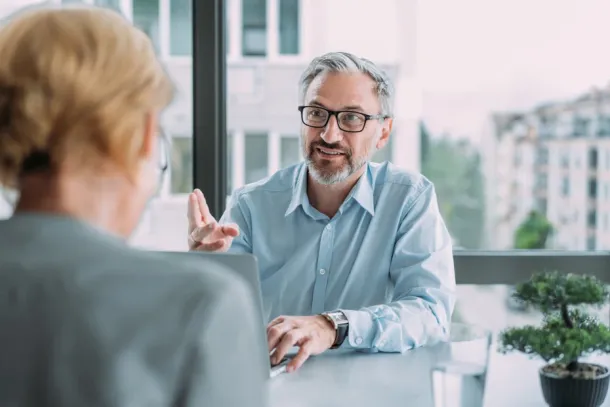 Shot of a two confident business persons sitting on a desk in the office and sharing ideas. Businessman and businesswoman in meeting using laptop and digital tablet discussing business strategy. Business coworkers working together in the office.