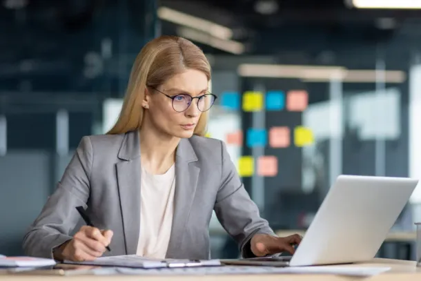 A professional businesswoman engages in serious work, meticulously analyzing paperwork and utilising her laptop.
