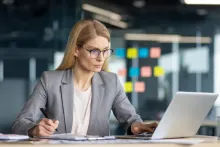 A professional businesswoman engages in serious work, meticulously analyzing paperwork and utilising her laptop.