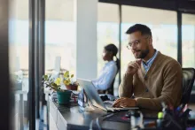 Young male design professional brainstorming while working on a computer in the office. His colleague is in the background.