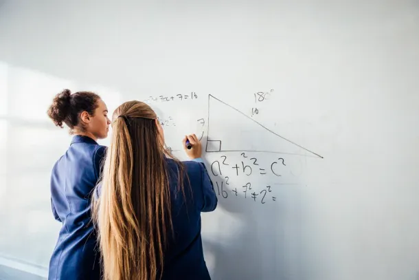 Two teenage school girls standing in front of a large whiteboard side by side solving a mathematics equation on the board. Back view