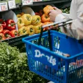 A person stands looking at vegetables in a supermarket, holding a blue basket.