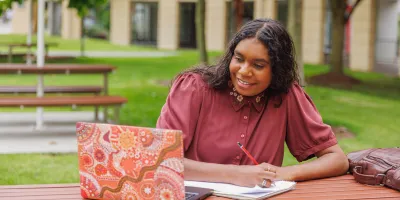 A smiling First Nations woman sits outside at a table writing in a notebook while looking at a laptop.