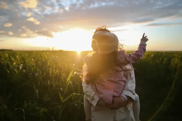 Women with a child on her back in a pink jacket. standing in a field with high grass and looking to the sunset pointing.