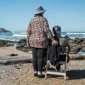 Two women on the shore looking at the ocean. One is sitting, one stands next her.